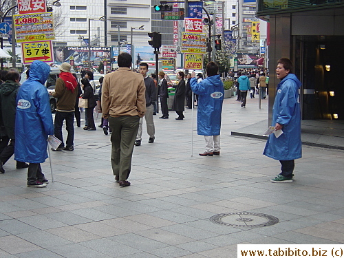 These men in blue coats give out fliers for a spectacles shop.  The sign the man on the left is holding says there're 57 steps to the shop. Not 58, just 57