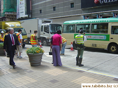 HK policeman. Minibus in the background. Wait a minute! Is HE on the cell phone too? No, it looks like a walkie talkie. Whew!
