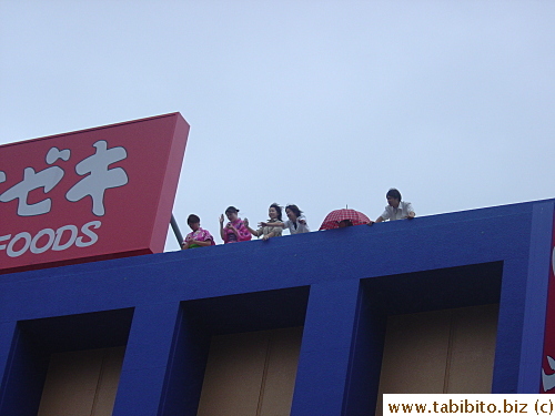 A few women watch the parade on roof top, two of them wear yukata
