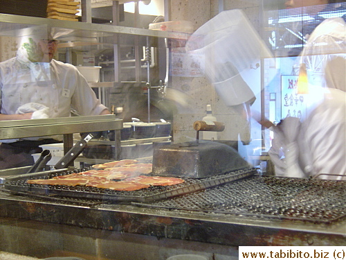 The chef grills thinly sliced meat and burger patties over charcoal behind a glass partition